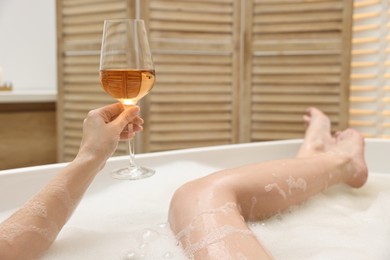 Photo of Woman with glass of wine taking bath in tub indoors, closeup