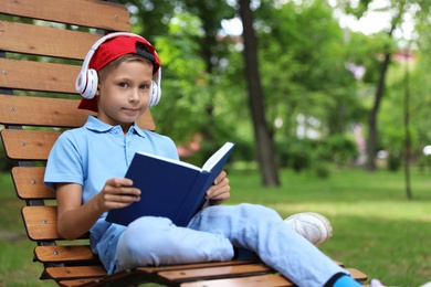 Cute little boy with headphones reading book on lounger in park