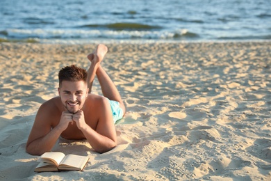 Young man reading book on sandy beach near sea