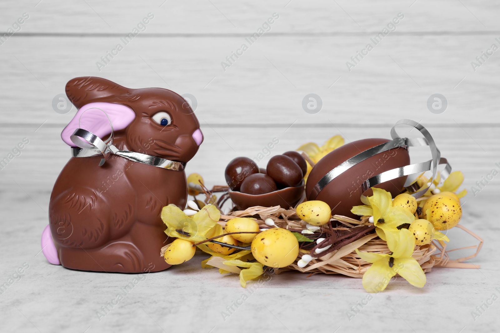 Photo of Easter celebration. Funny chocolate bunny near nest with sweet eggs and candies on white textured background