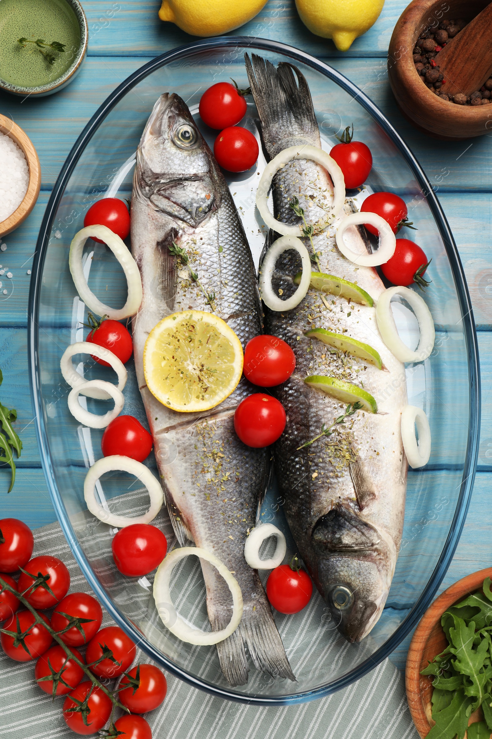 Photo of Glass baking tray with sea bass fish and ingredients on light blue wooden table, flat lay