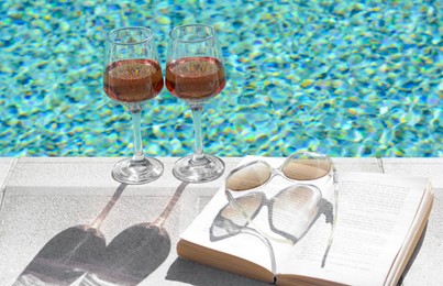 Glasses of tasty rose wine, open book and sunglasses on swimming pool edge