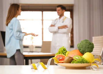 Photo of Nutritionist consulting patient at table in clinic, focus on plate with fruits, vegetables and measuring tape