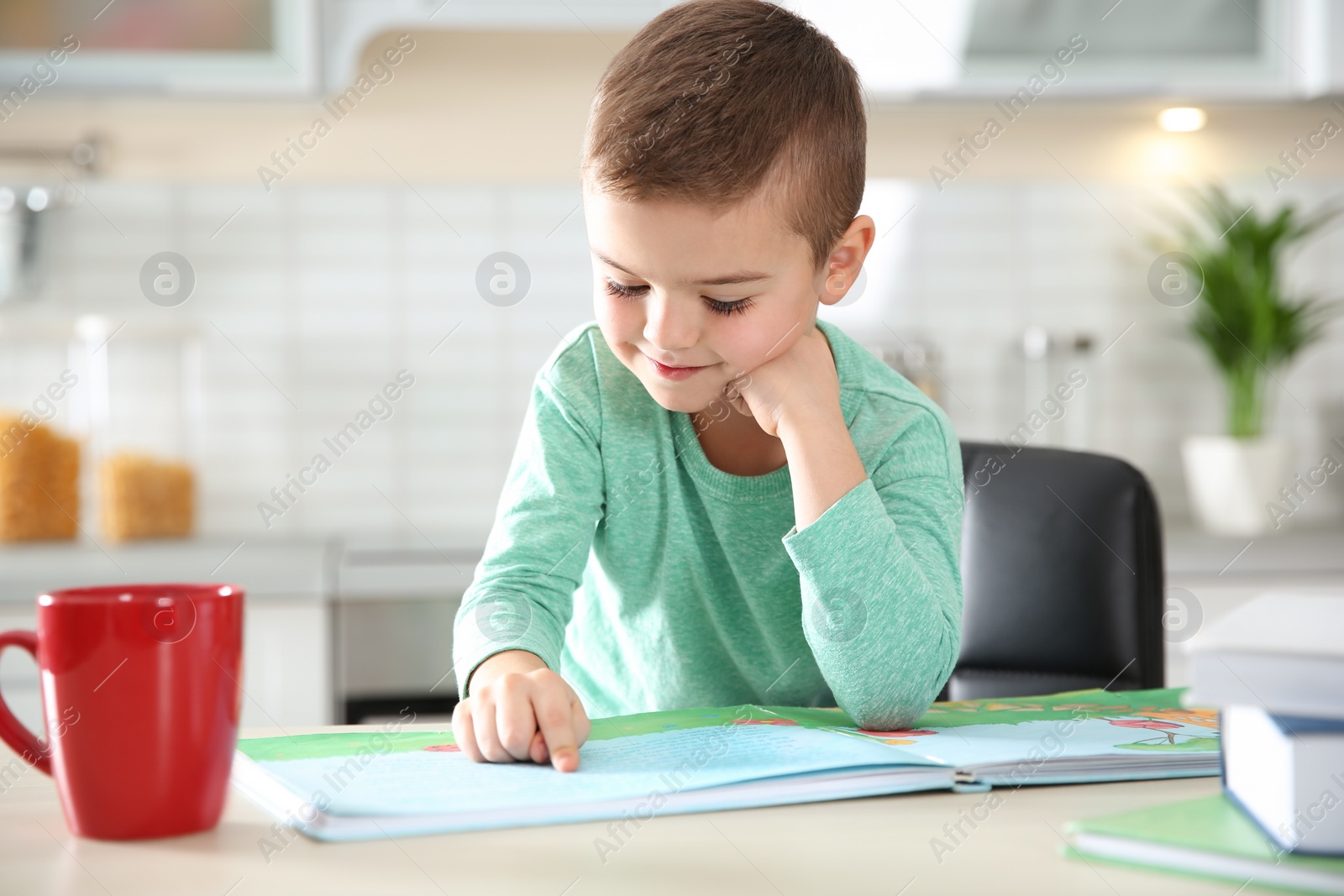 Photo of Cute little boy reading book at table in kitchen