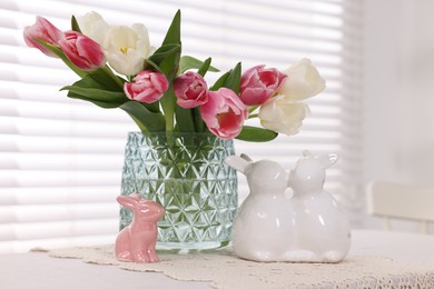 Photo of Easter decorations. Bouquet of tulips in vase and bunny figures on white table at home
