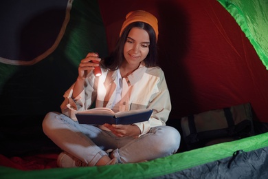 Young woman with flashlight reading book in tent at night