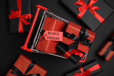 Photo of Small shopping cart with wrapped gift boxes on dark background, flat lay. Black Friday sale