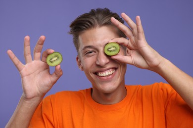 Smiling man covering eye with half of kiwi on violet background
