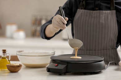 Photo of Woman cooking delicious crepe on electric pancake maker at white marble table in kitchen, closeup