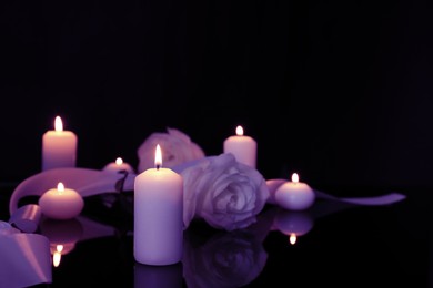 Violet roses and burning candles on black mirror surface in darkness, space for text. Funeral attributes