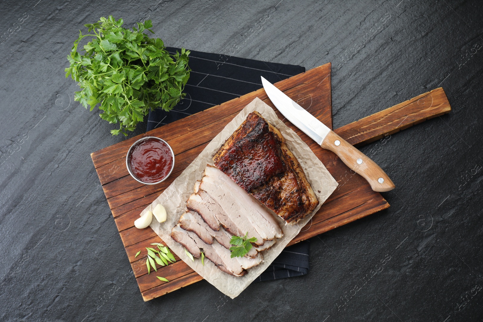 Photo of Pieces of baked pork belly served with sauce and parsley on black textured table, top view