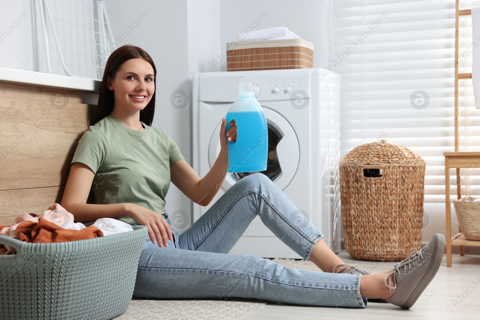 Photo of Woman sitting on floor near washing machine and holding fabric softener in bathroom