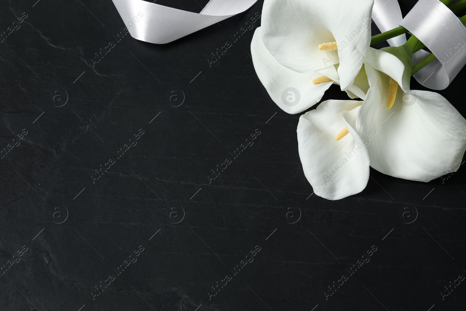 Photo of Beautiful calla lily flowers and white ribbon on black table, above view with space for text. Funeral symbols