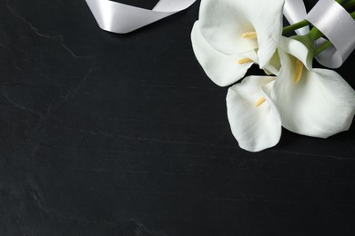 Photo of Beautiful calla lily flowers and white ribbon on black table, above view with space for text. Funeral symbols