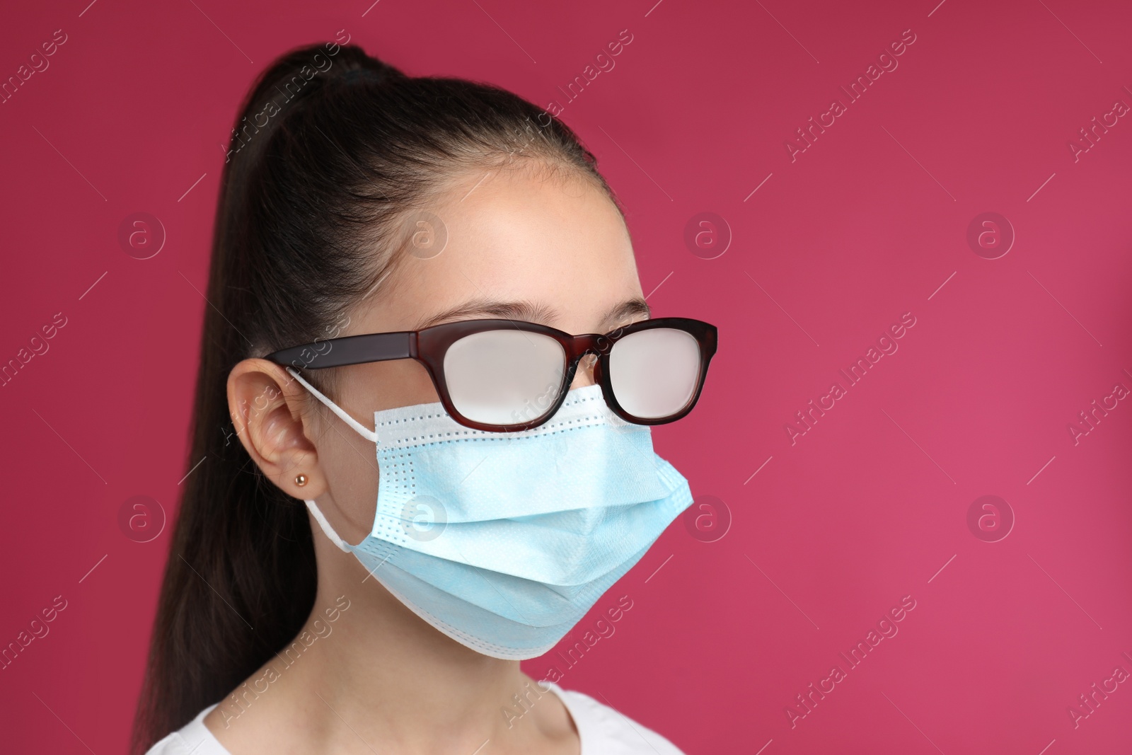 Photo of Little girl with foggy glasses caused by wearing medical face mask on pink background. Protective measure during coronavirus pandemic