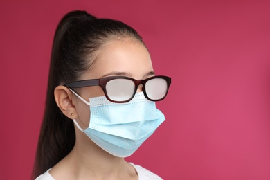 Little girl with foggy glasses caused by wearing medical face mask on pink background. Protective measure during coronavirus pandemic