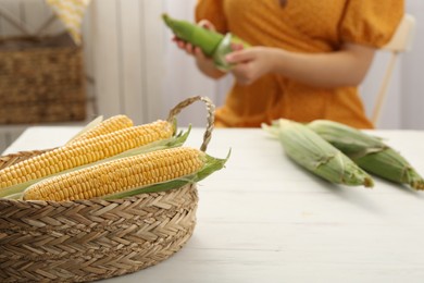 Photo of Woman husking corn cob at white wooden table, focus on basket