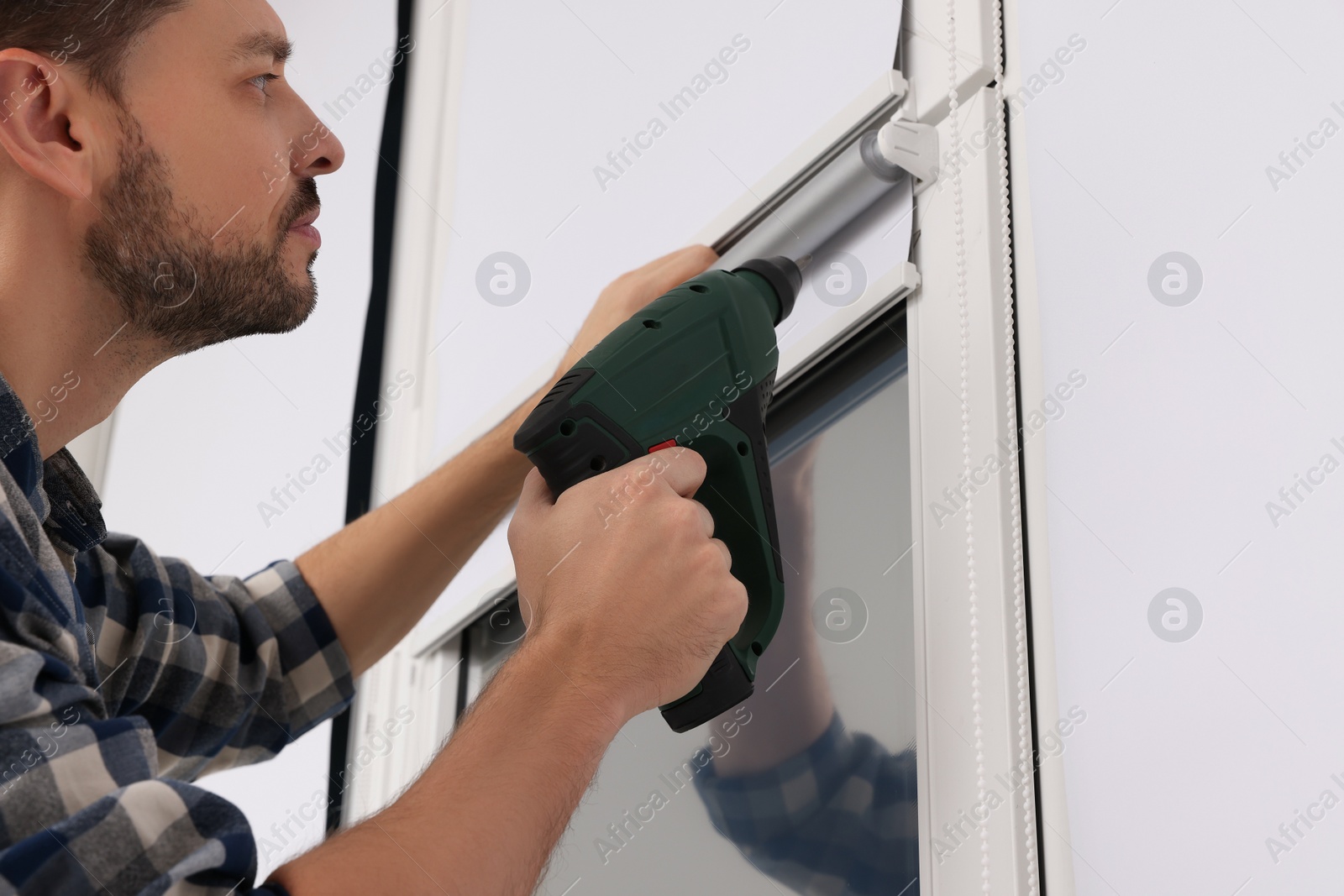 Photo of Man with drill installing roller window blind indoors, closeup