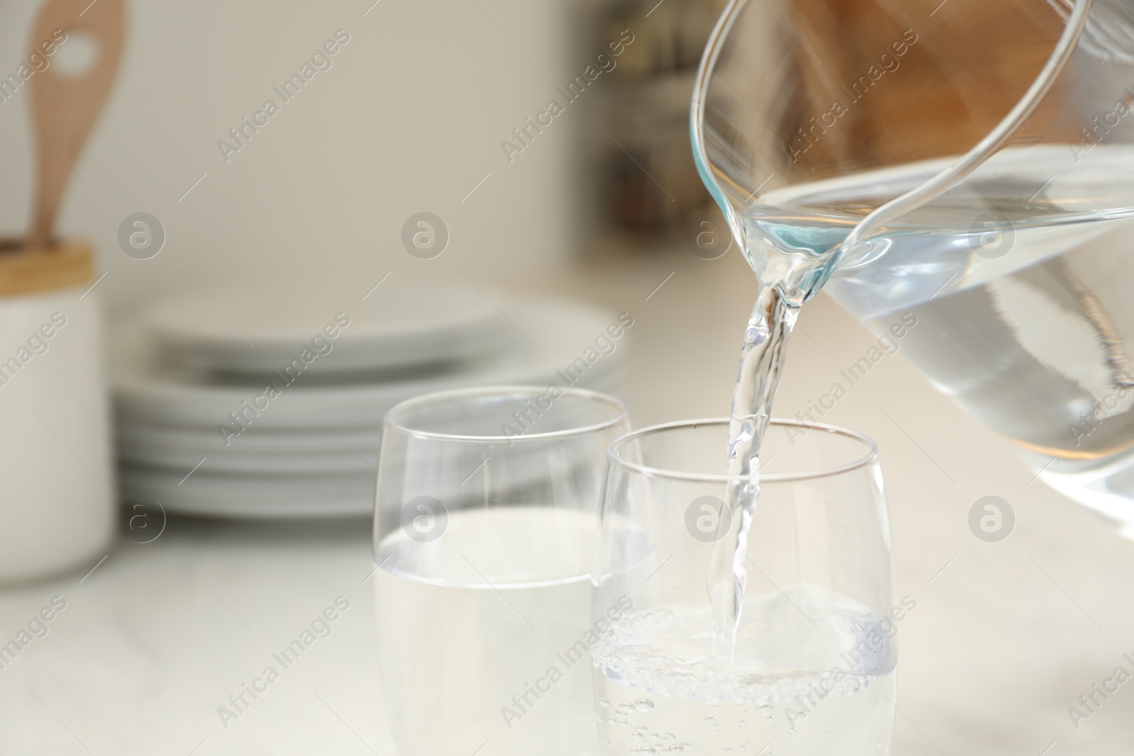 Photo of Pouring water from jug into glass at white table in kitchen, closeup