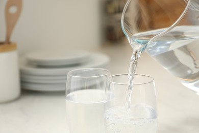 Pouring water from jug into glass at white table in kitchen, closeup