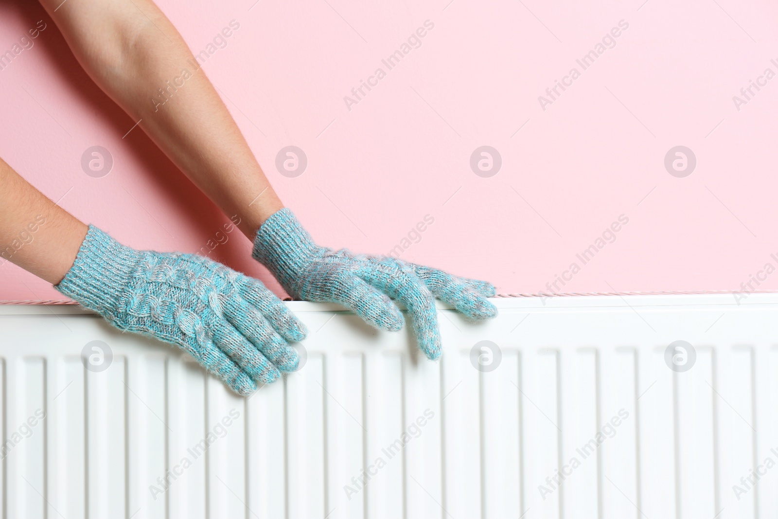 Photo of Woman in gloves warming hands on heating radiator near color wall. Space for text