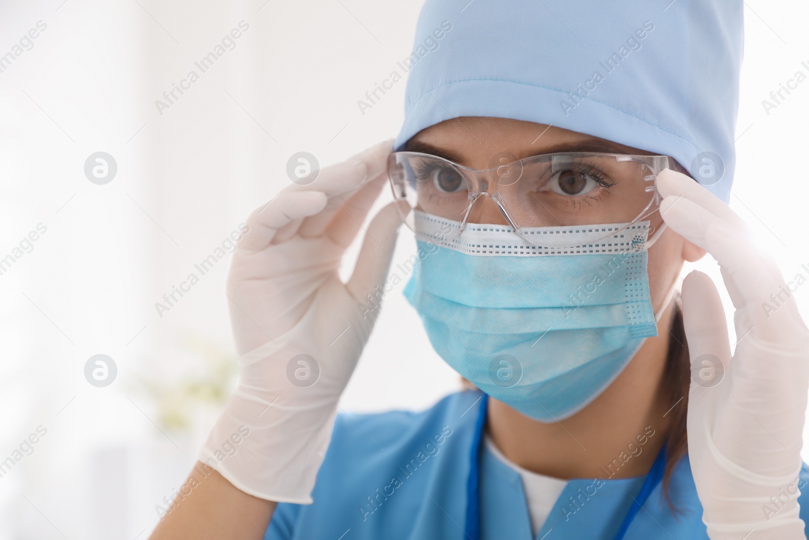 Photo of Doctor in medical gloves and protective mask putting on glasses against light background, closeup