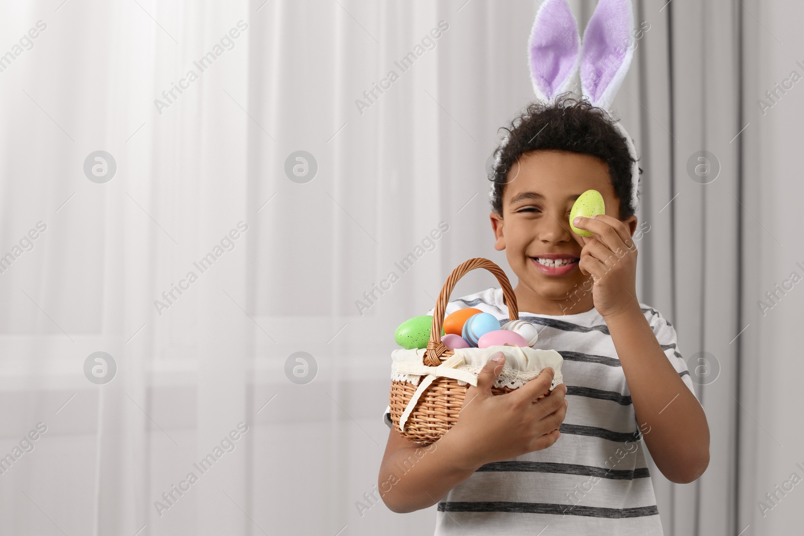 Photo of Cute African American boy in bunny ears headband covering eye with Easter egg indoors, space for text