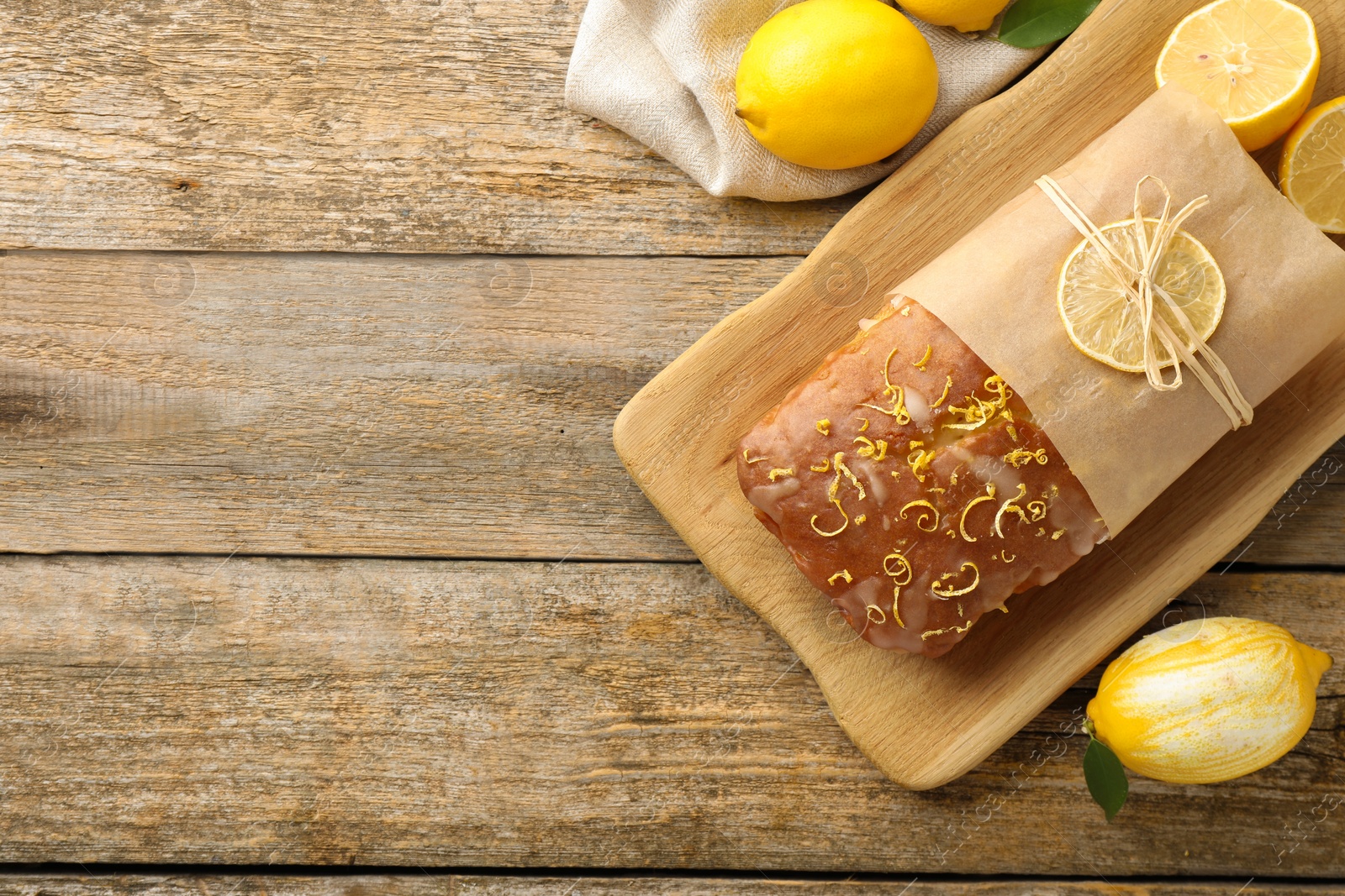 Photo of Wrapped tasty lemon cake with glaze and citrus fruits on wooden table, flat lay. Space for text