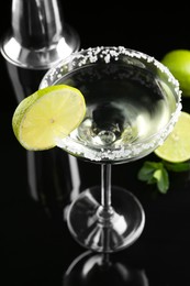 Photo of Delicious cocktail with salt, lime and metal shaker on black mirror surface, closeup