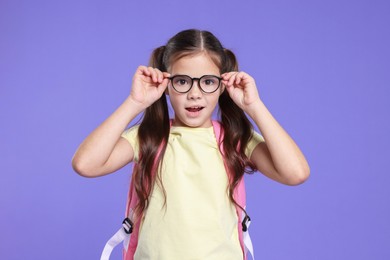Photo of Cute schoolgirl in glasses with backpack on violet background
