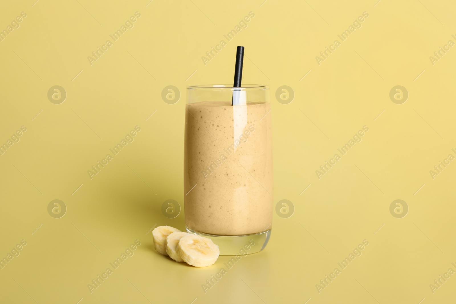 Photo of Glass of tasty smoothie with straw and cut banana on pale yellow background