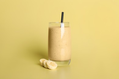 Glass of tasty smoothie with straw and cut banana on pale yellow background