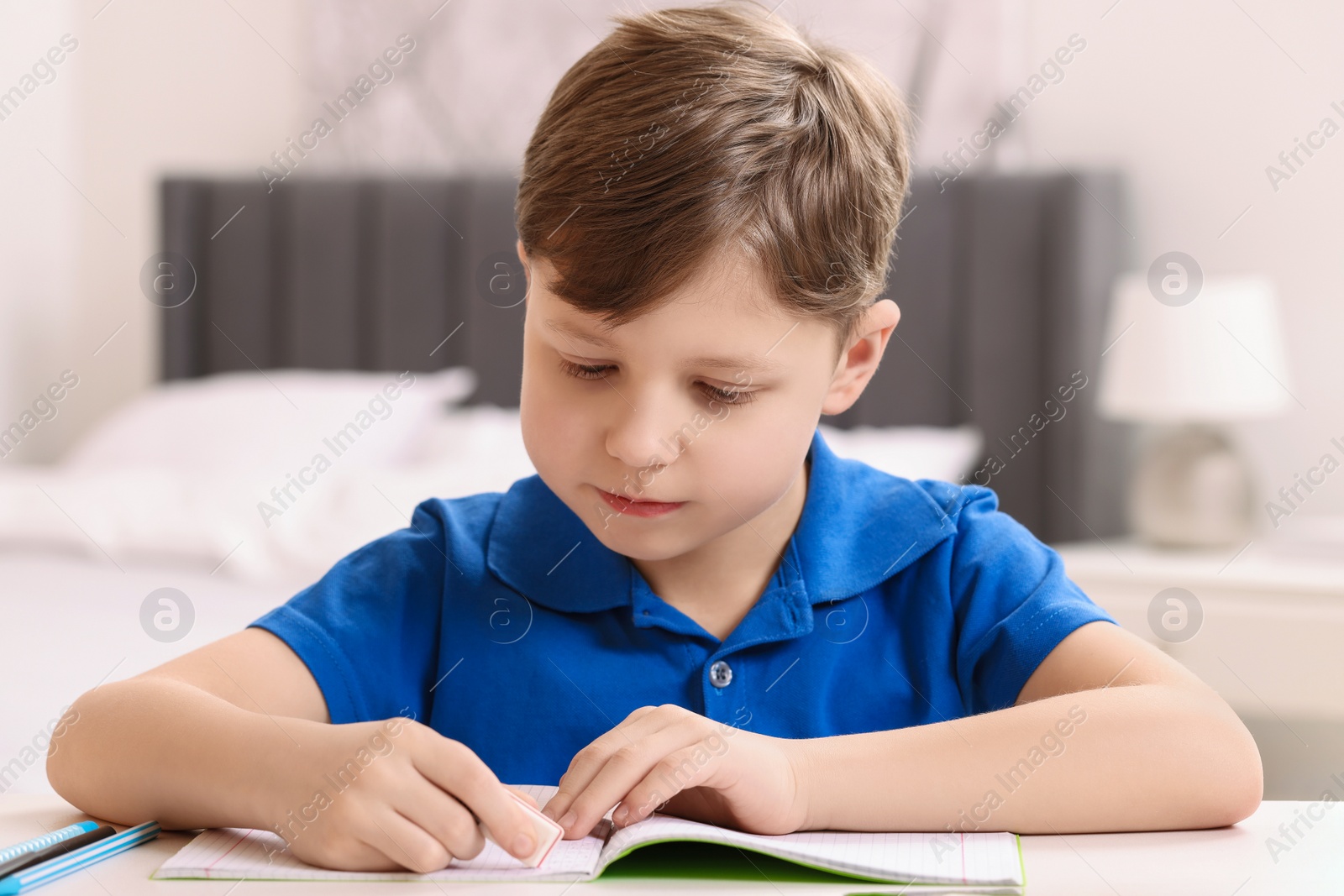 Photo of Little boy erasing mistake in his notebook at white desk indoors