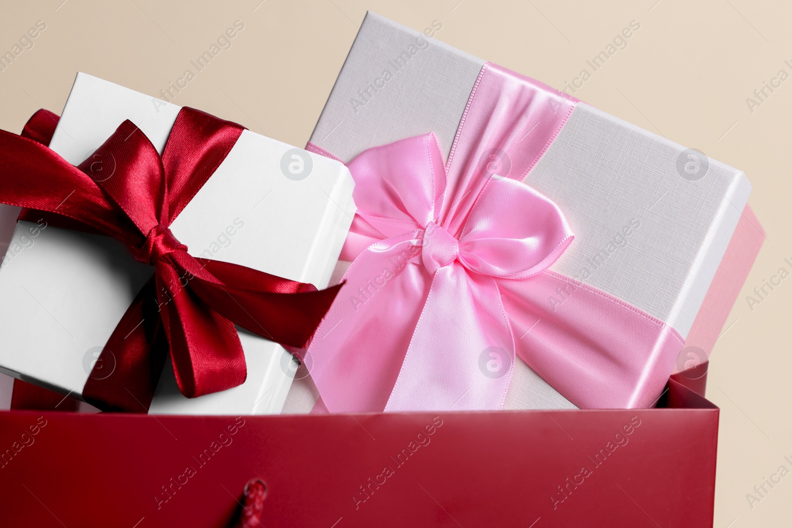 Photo of Dark red paper shopping bag full of gift boxes on beige background, closeup