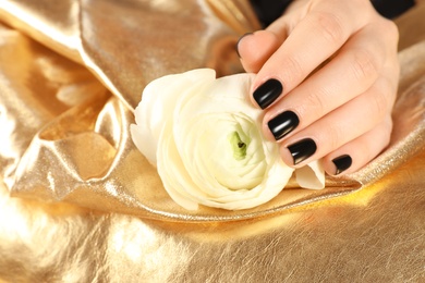 Photo of Woman with black manicure holding beautiful flower on golden fabric, closeup. Nail polish trends