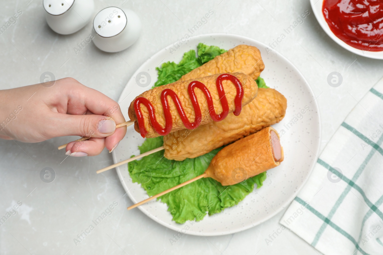Photo of Woman holding delicious corn dog with ketchup at light table, top view