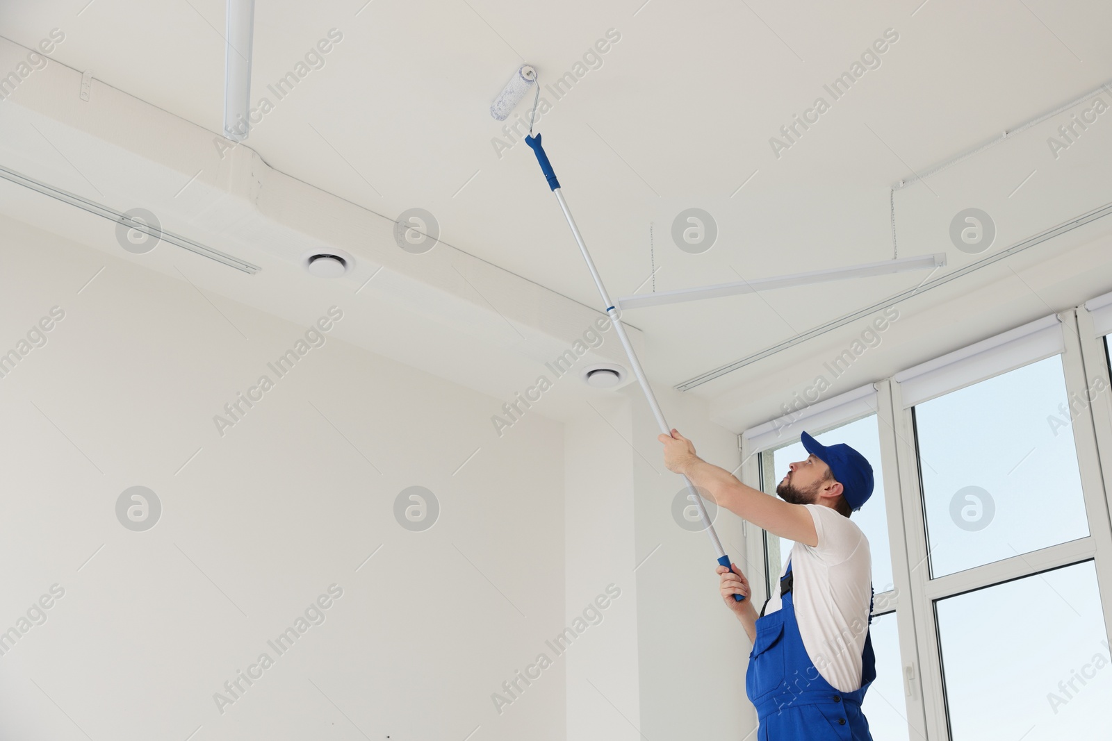 Photo of Worker in uniform painting ceiling with roller indoors. Space for text