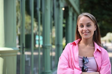 Beautiful young woman with stylish sunglasses near building outdoors, space for text