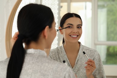 Photo of Beautiful young woman applying mascara in front of mirror at home
