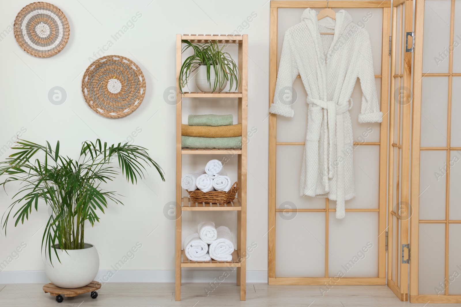Photo of Soft folded towels on wooden shelving unit and bathrobe near white wall
