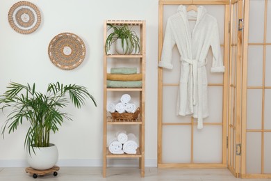 Photo of Soft folded towels on wooden shelving unit and bathrobe near white wall