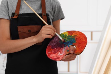 Woman mixing paints on palette with brush near easel in studio, closeup