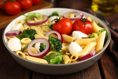 Photo of Bowl of delicious pasta with tomatoes, onion and broccoli on wooden table, closeup