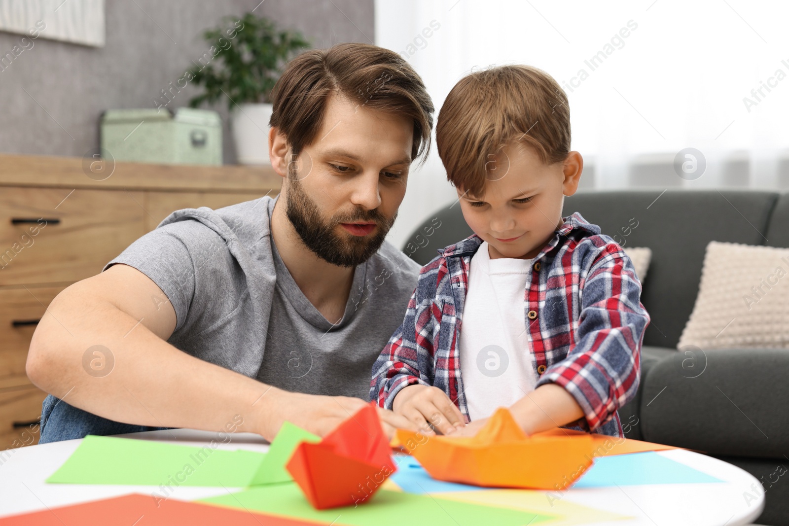 Photo of Dad and son making paper boats at coffee table indoors