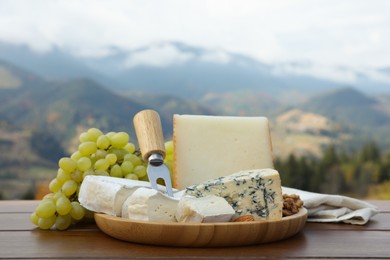 Different types of delicious cheeses, nuts and grapes on wooden table against mountain landscape