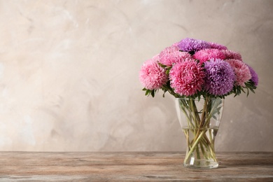 Photo of Glass vase with beautiful aster flowers on wooden table against beige background. Space for text