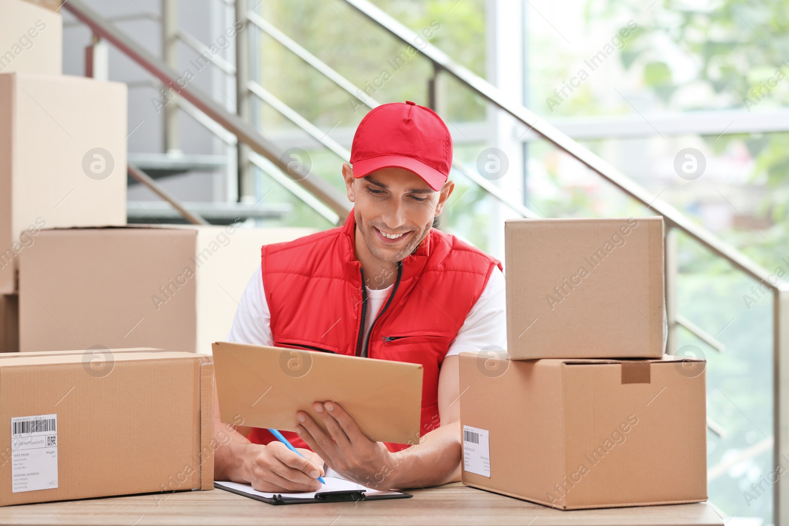 Photo of Young courier working with papers among parcels at table in delivery department