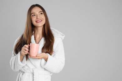 Photo of Beautiful young woman in bathrobe with cup of morning drink on light grey background, space for text