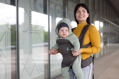 Mother holding her child in sling (baby carrier) near building outdoors