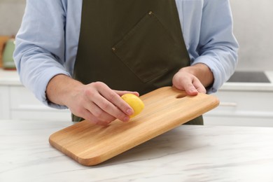 Photo of Man rubbing wooden cutting board with lemon at white table in kitchen, closeup